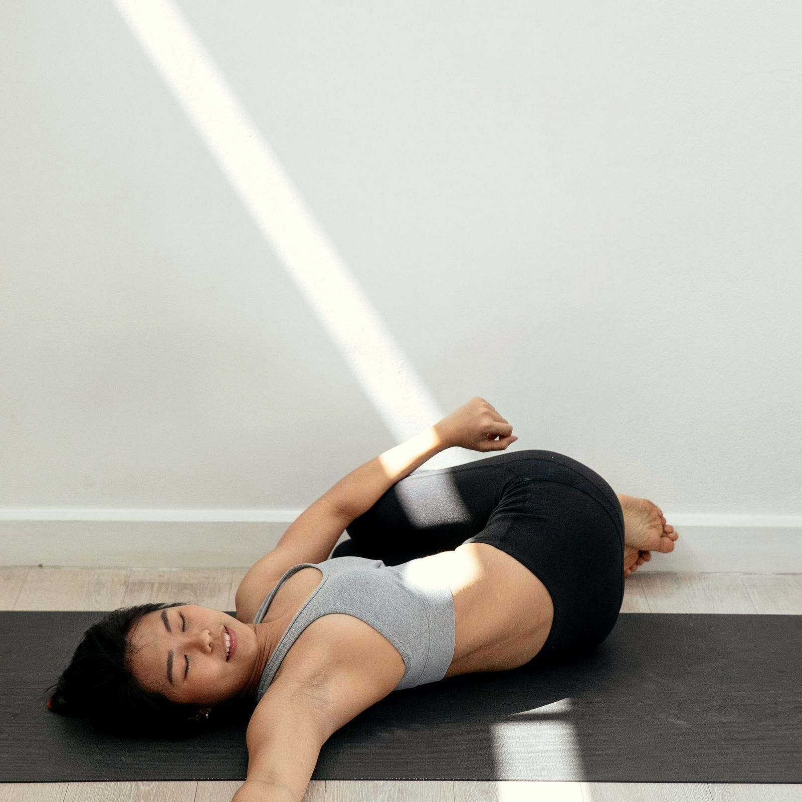 An Asian woman in a reclining twist in a sharp band of sunlight by a white wall