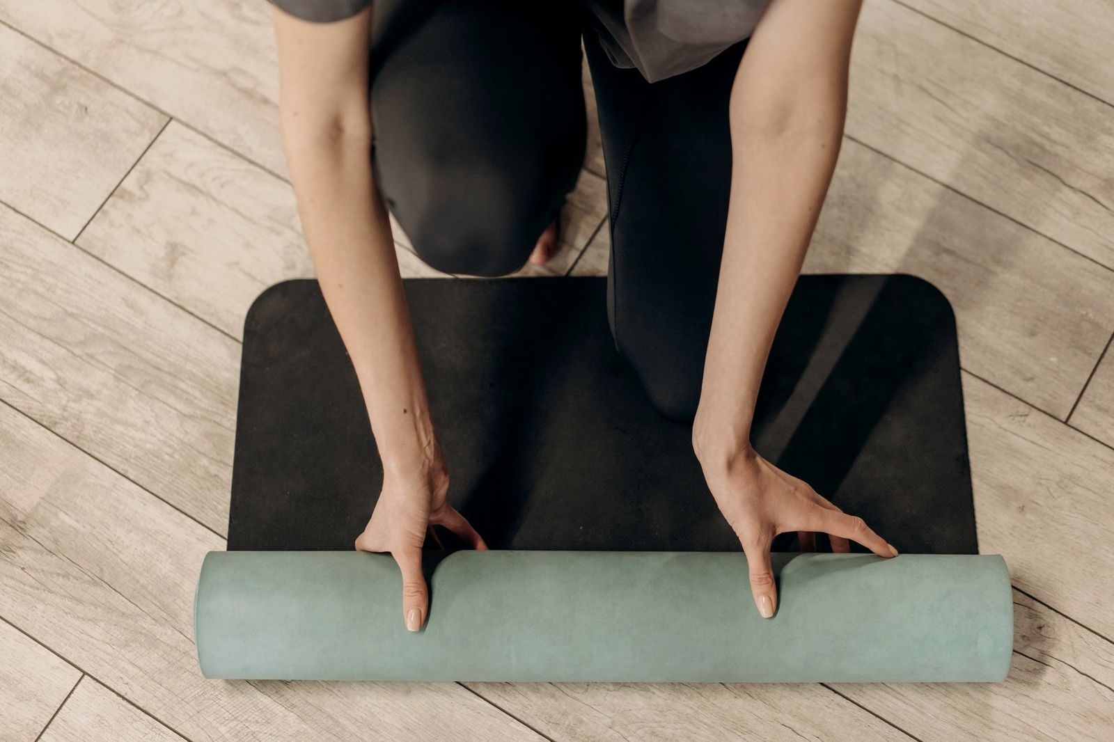 Hands unrolling a sage-green yoga mat on a wooden floor, seen from above