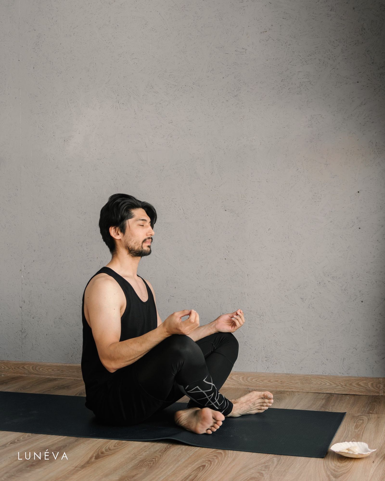 A man in a black activewear set seated on a yoga mat in a quiet, bright room